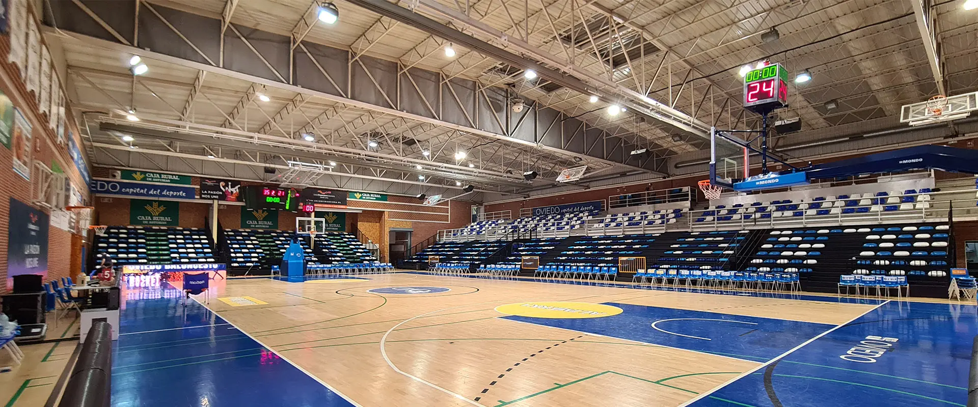 Pista polideportiva, zonas laterales en azul, círculos amarillos. Gradas, dos tableros, canasta y aro de baloncesto, marcador
