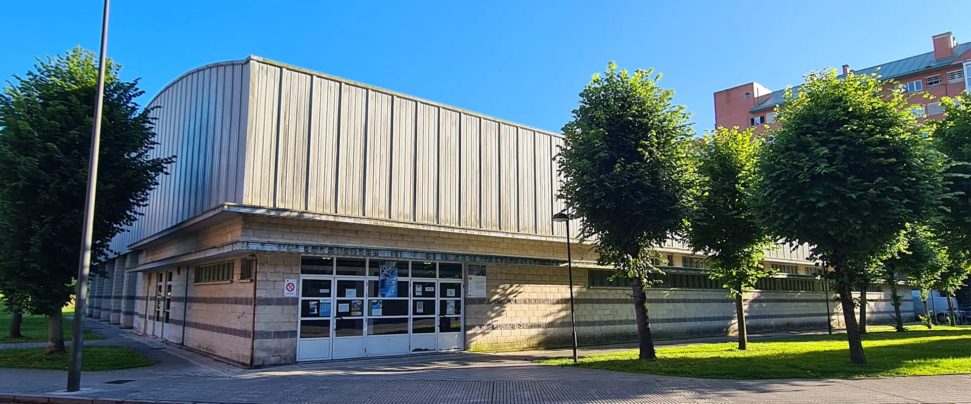 Lateral del edificio de hormigón y techo metálico. Destaca la puerta principal blanca con rectángulos de cristal  
