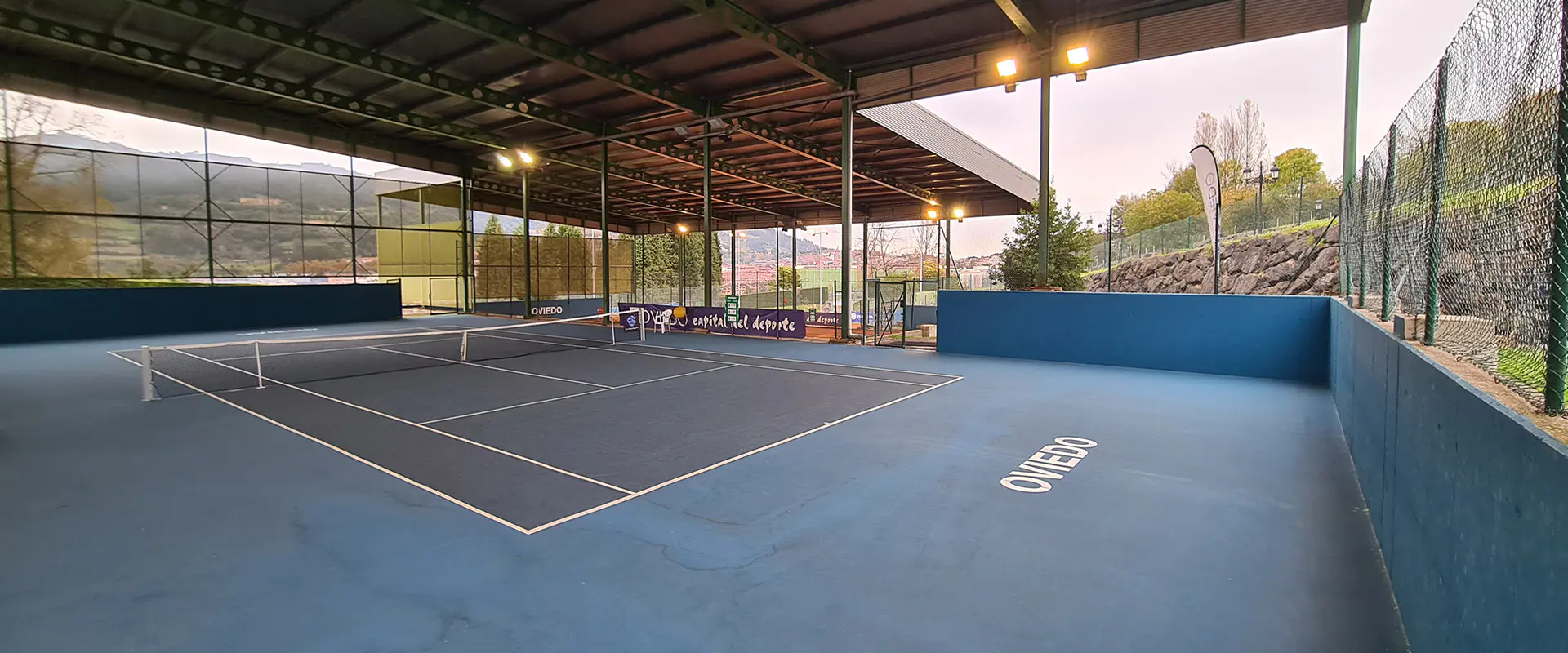 Vista a pie de pista de una cancha de tenis cubierta. Muros y vallas perimetrales. Puertas de acceso