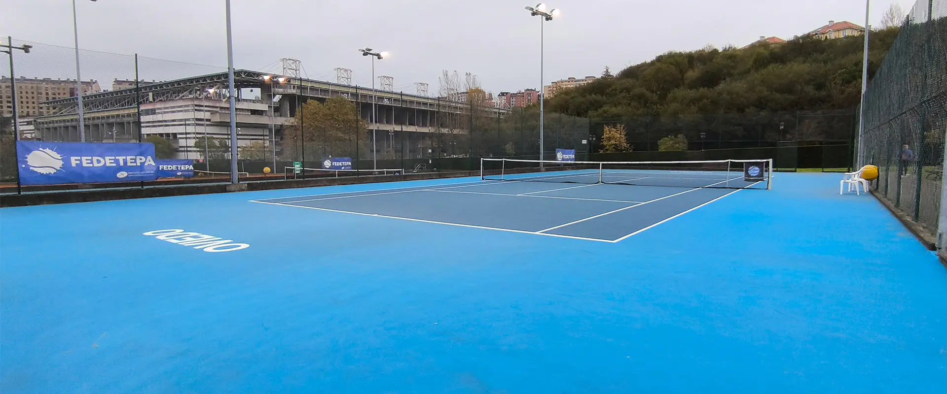 Panorámica de una pista de tenis descubierta, con vallas perimetrales y el Estadio Carlos Tartiere en un lateral