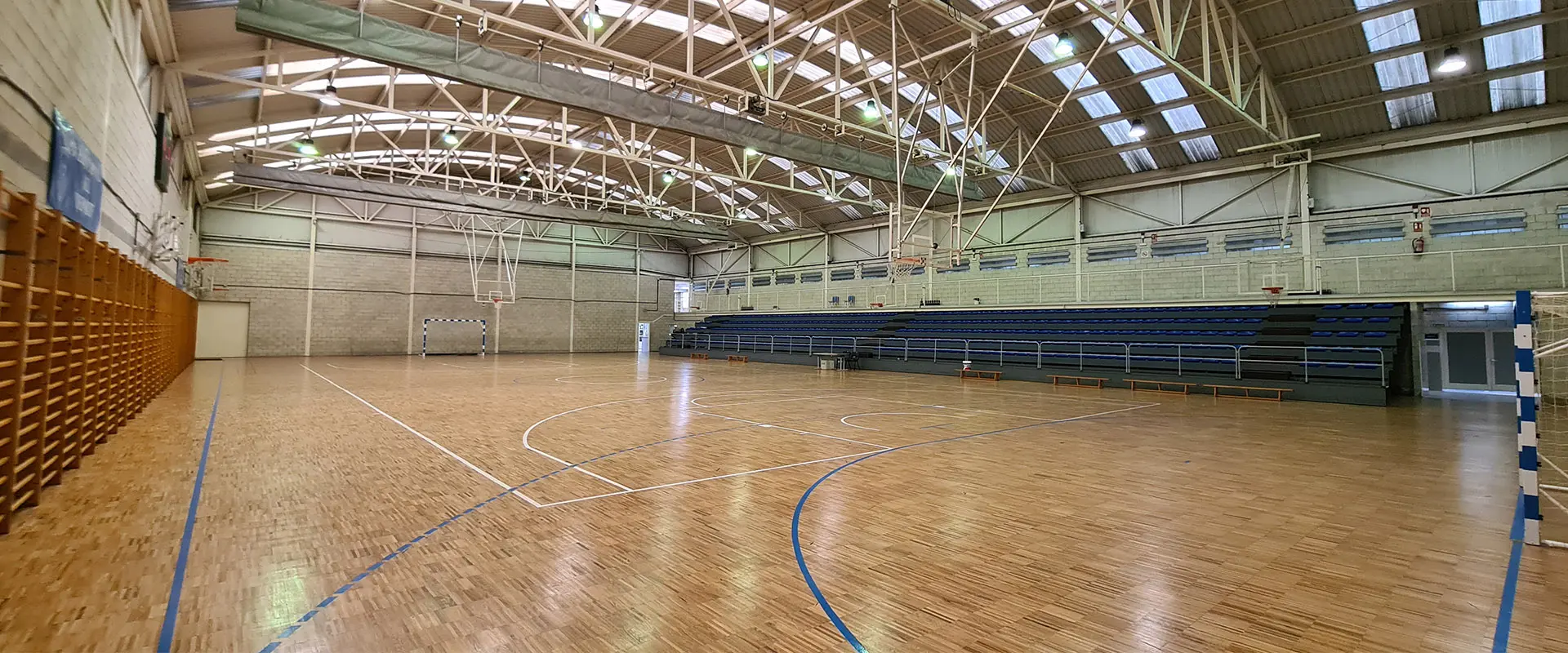 Pista polideportiva de parquet y grada tomada a pie de pista, desde uno de los córneres. Los asientos de las grada son azules