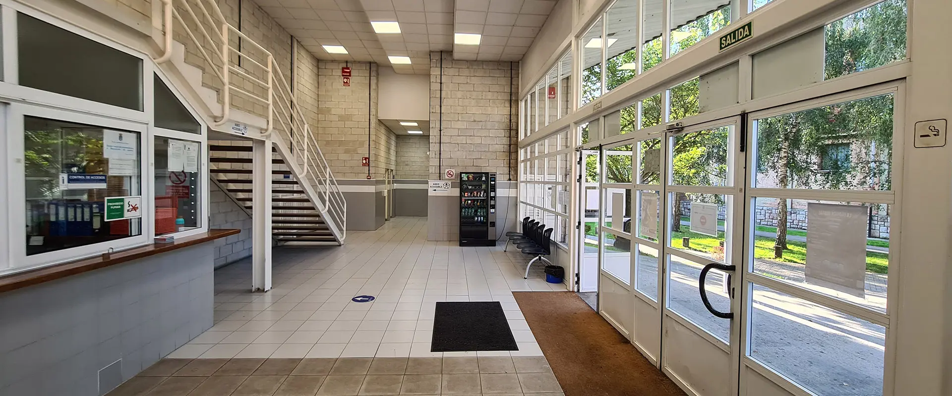 Hall de entrada instalación con amplio ventanal y puertas.  Mostrador acristalado de recepción y escalera al primer piso. 