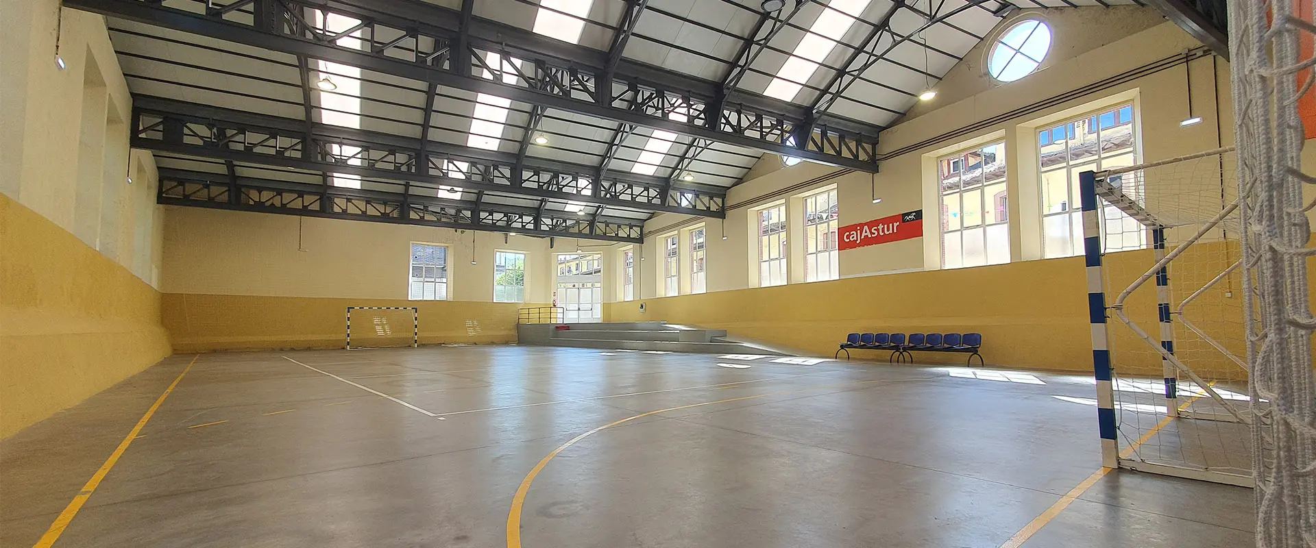 Pista polideportiva tomada desde el área del córner. Fondo zona público y puerta de entrada . Amplios ventanales