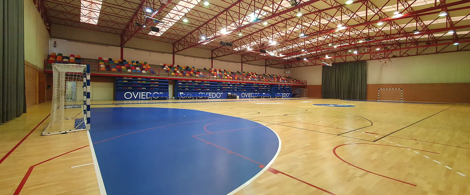 Pista polideportiva, tomada desde la pista, costado de portería. Gradas extensibles cerradas y  grada superior de colores