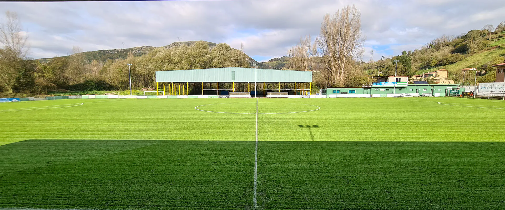 Vista frontal a pie de campo del terreno de juego, con instalaciones deportivas y edificios de servicios al fondo