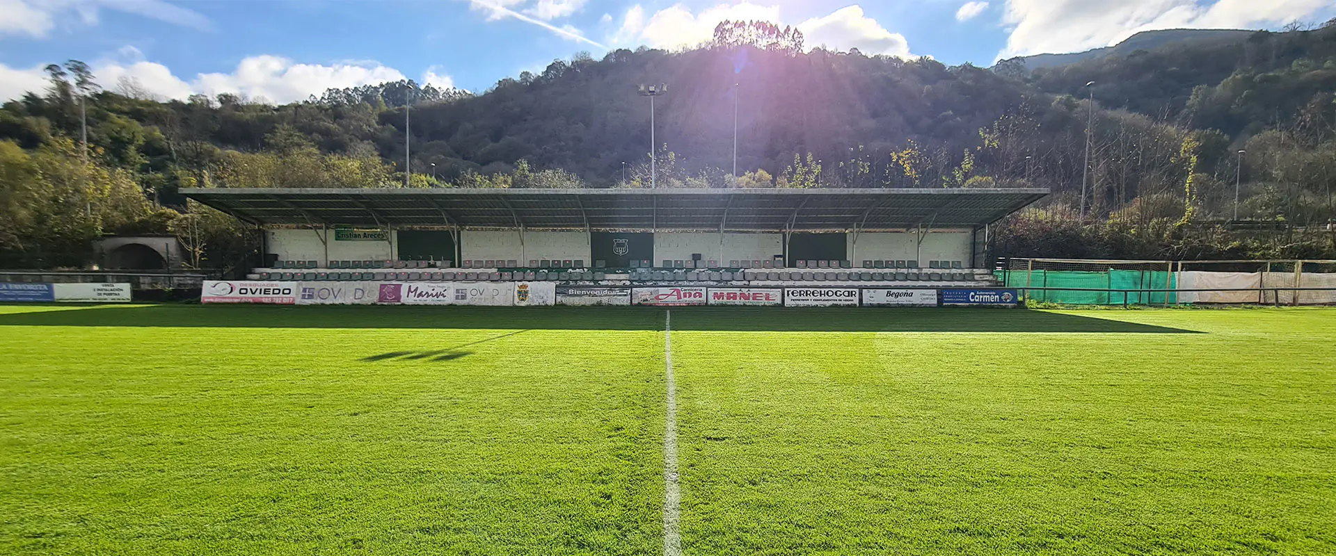 Vista frontal a pie de campo del terreno de juego, con las gradas al otro lado y un fondo de montañas