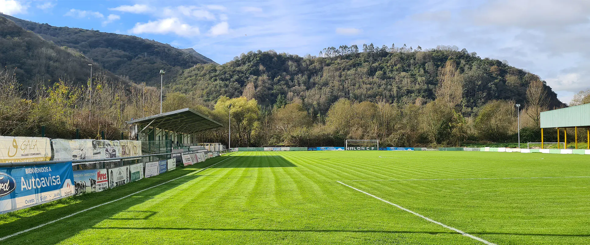 Campo desde una de las bandas, con gradas y vallas publicitarias a un lado, instalaciones al otro y una portería al fondo