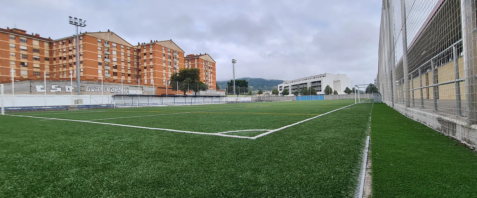 Terreno de juego a pie de campo y desde un córner, con vista de las vallas laterales y edificios circundantes al fondo