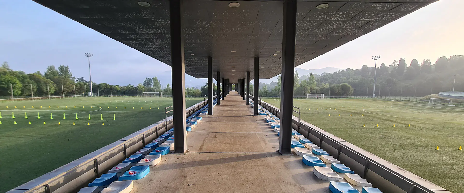 Pasillo cubierto de acceso a las gradas, con columnas. Vista parcial de los dos campos de fútbol 11 a los lados