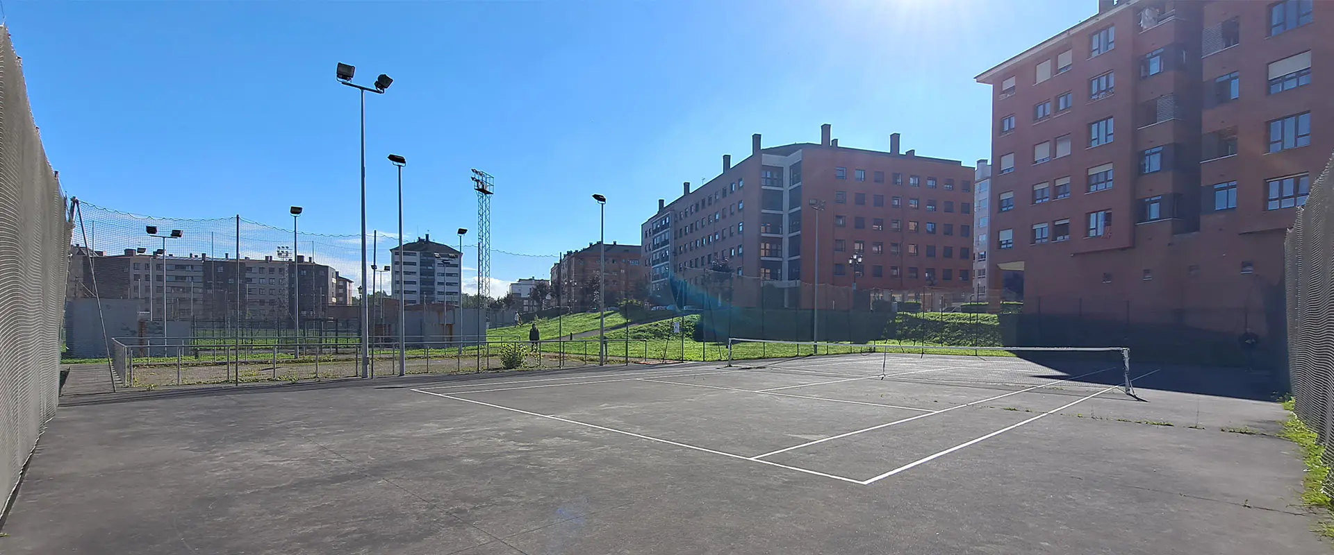 Vista lateral de una de las pistas de tenis, con torres de iluminación y edificios de viviendas al fondo