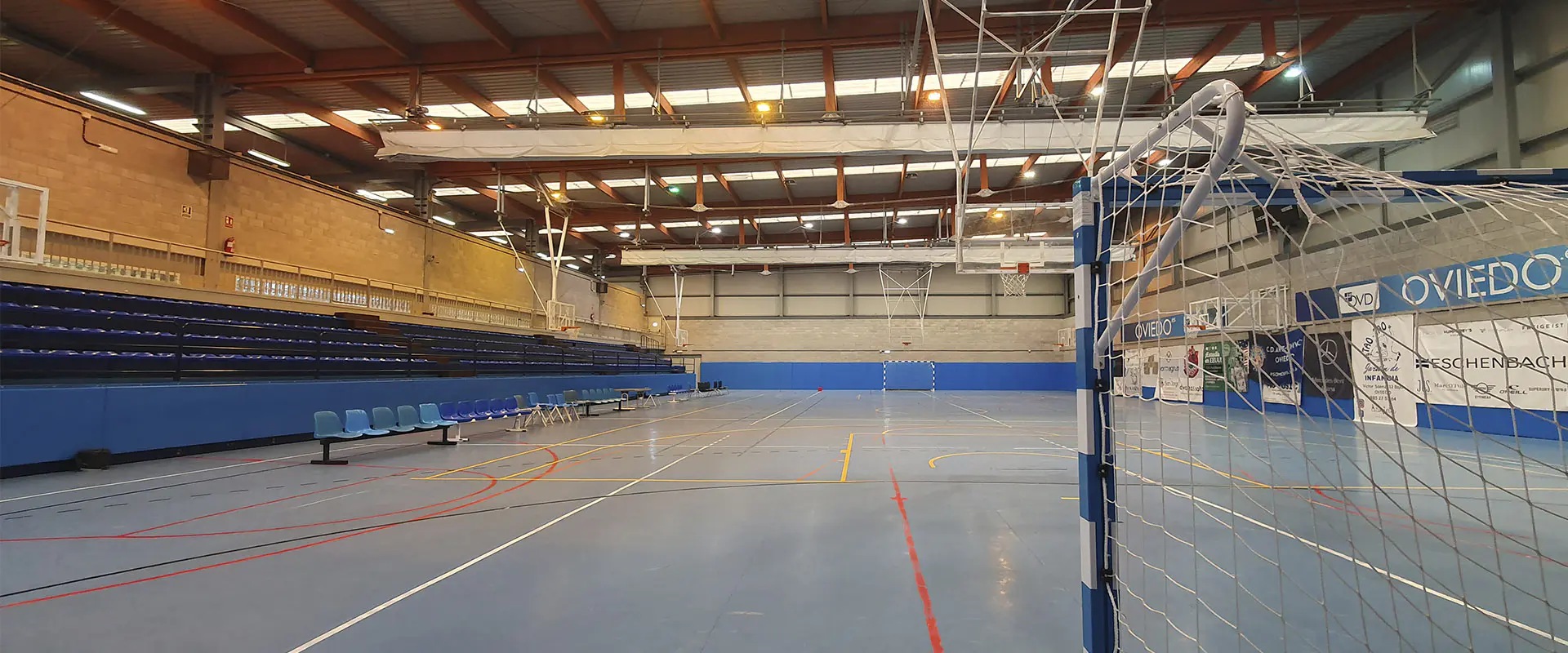 Pista polideportiva, fondo y lado izquierdo grada con asientos azules. Imagen tomada desde detrás de portería de fútbol 