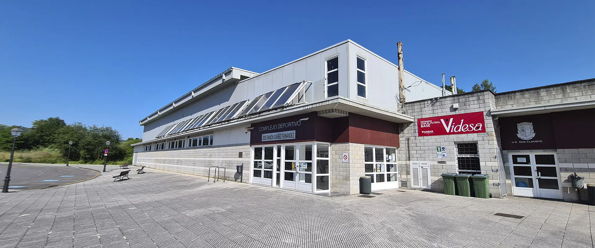 Exterior del polideportivo. Puerta principal metálica blanca con rectángulos de cristal. Hilera placas solares sobre alero