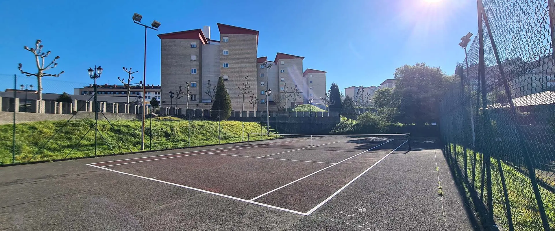Panorámica a pie de pista y desde una esquina de una cancha de tenis, con las vallas perimetrales y edificios al fondo