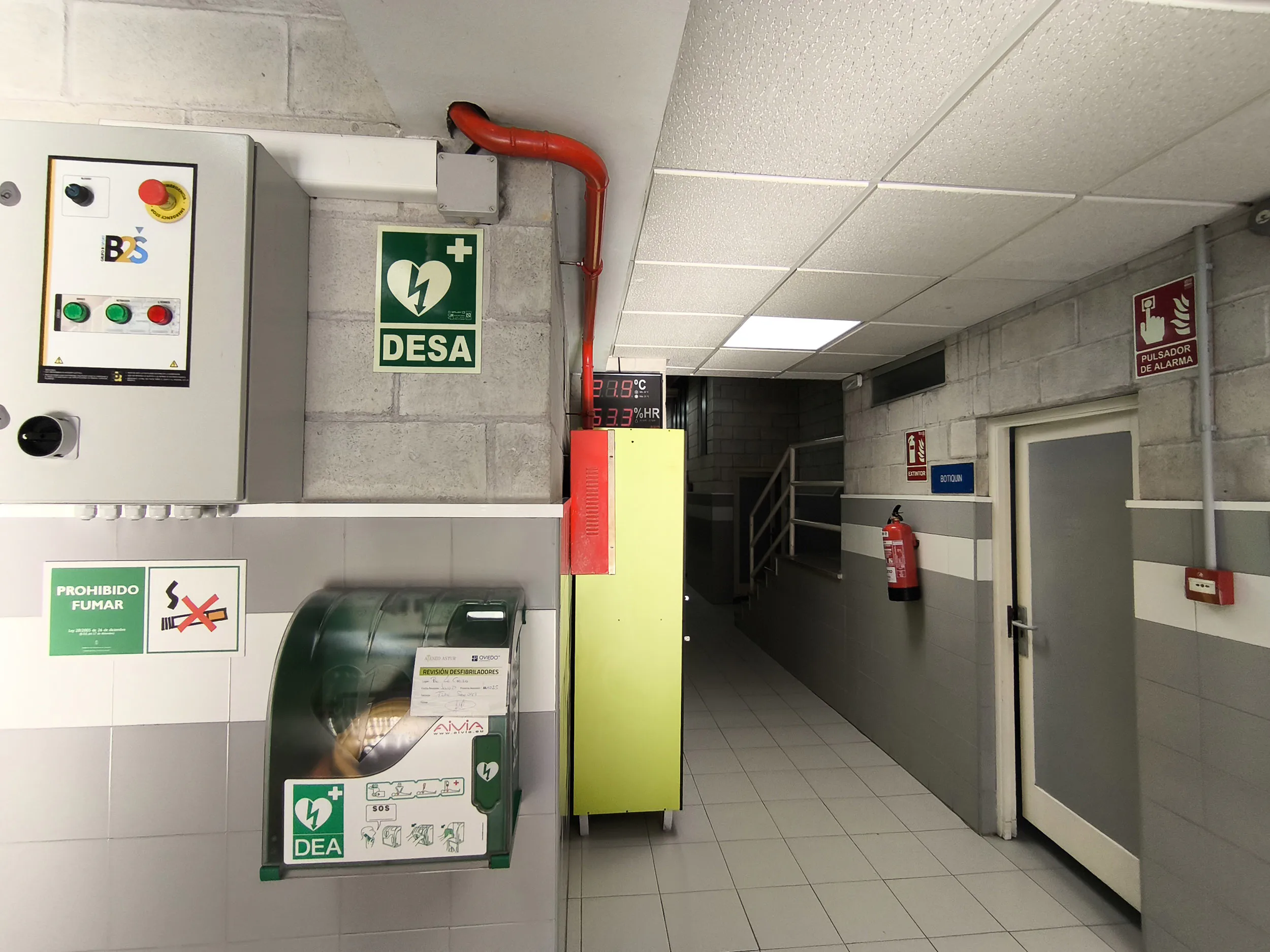 Pasillo de acceso a vestuarios y planta superior. Armario de maniobras eléctricas y aparato desfibrilador semiautomático