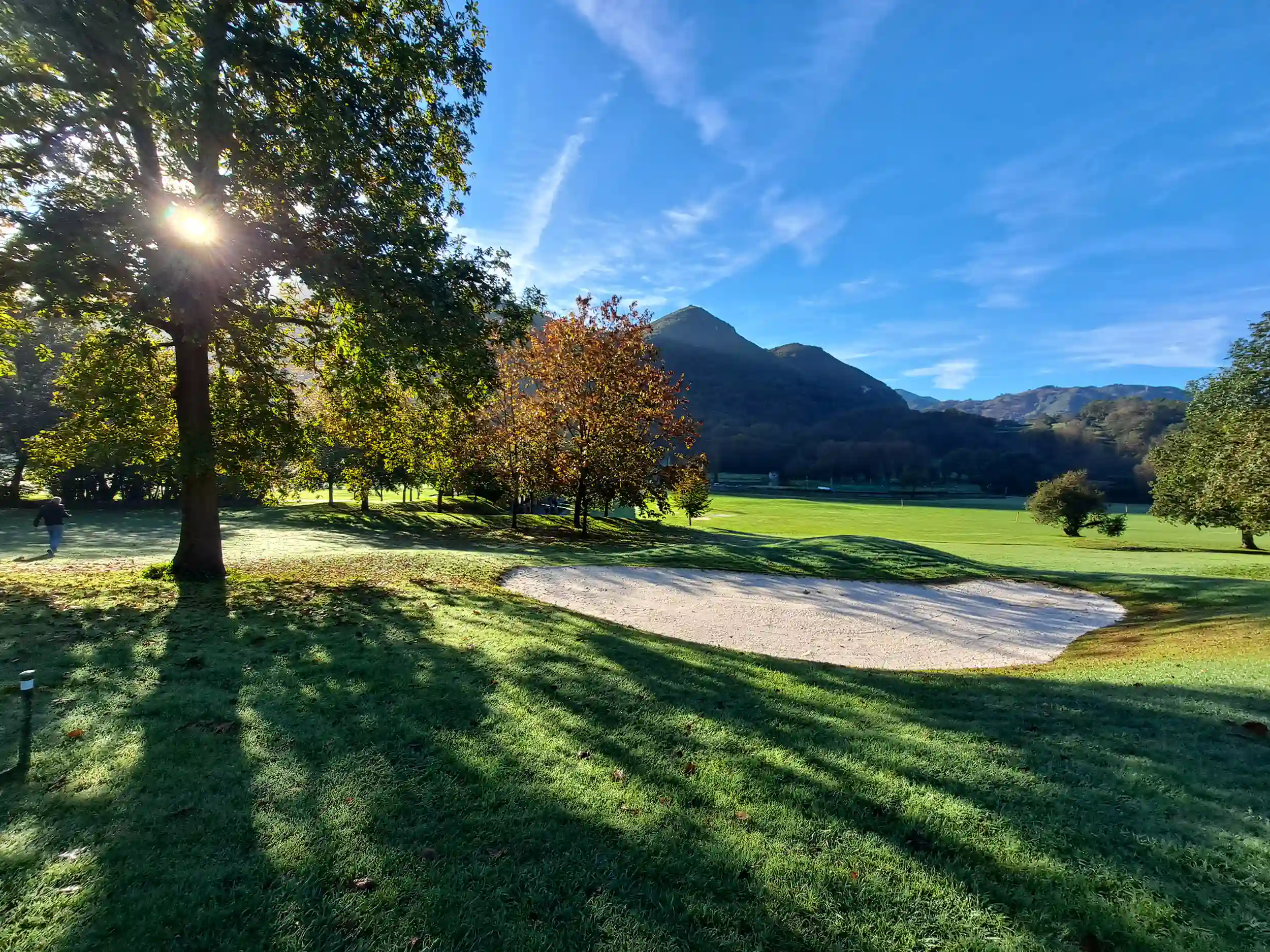 Panorámica de una zona del campo de golf, con un búnker de arena, árboles y un fondo montañoso