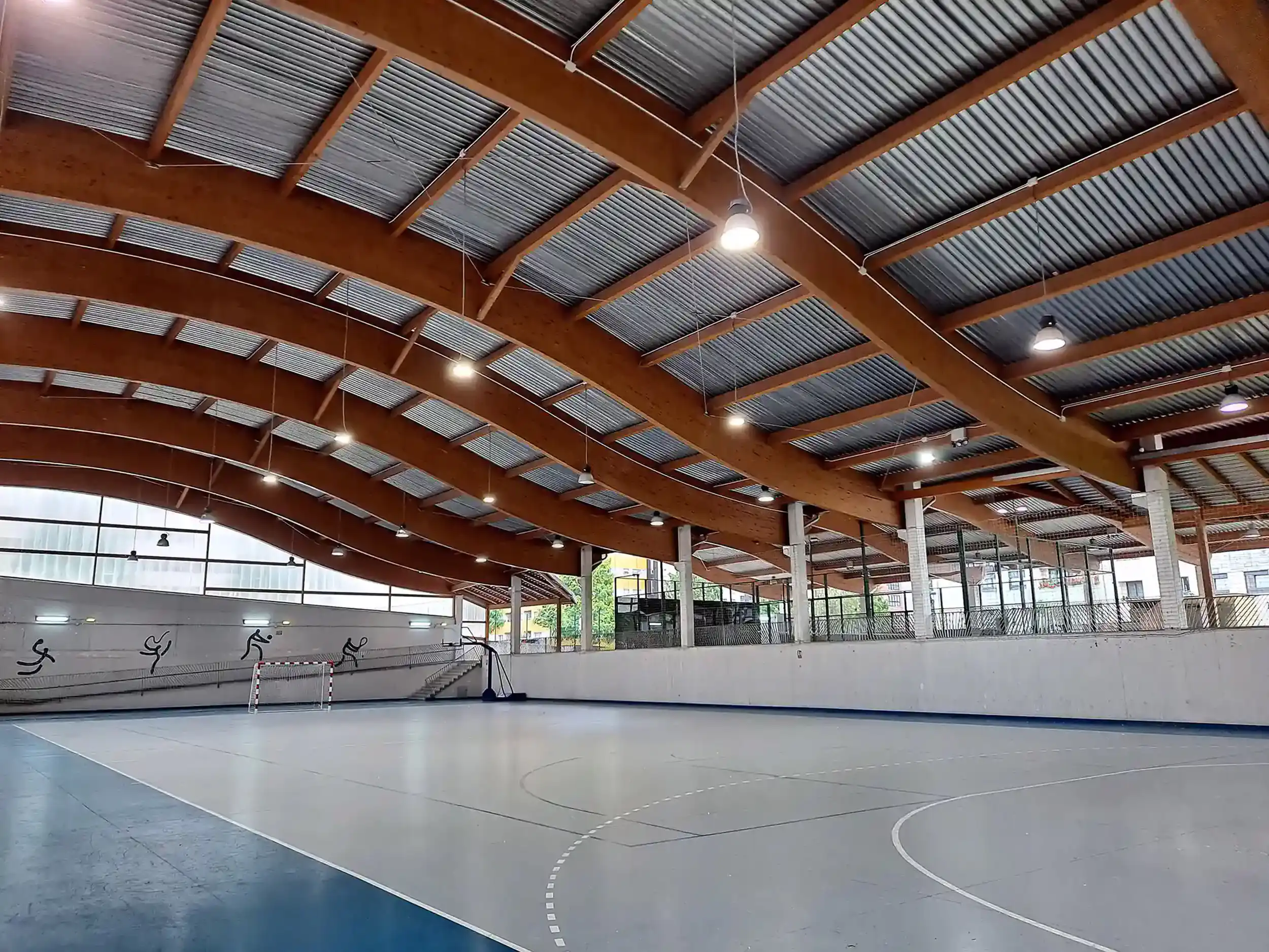Vista de la pista polideportiva, con portería y canasta al fondo y una cubierta ondulada de vigas de madera