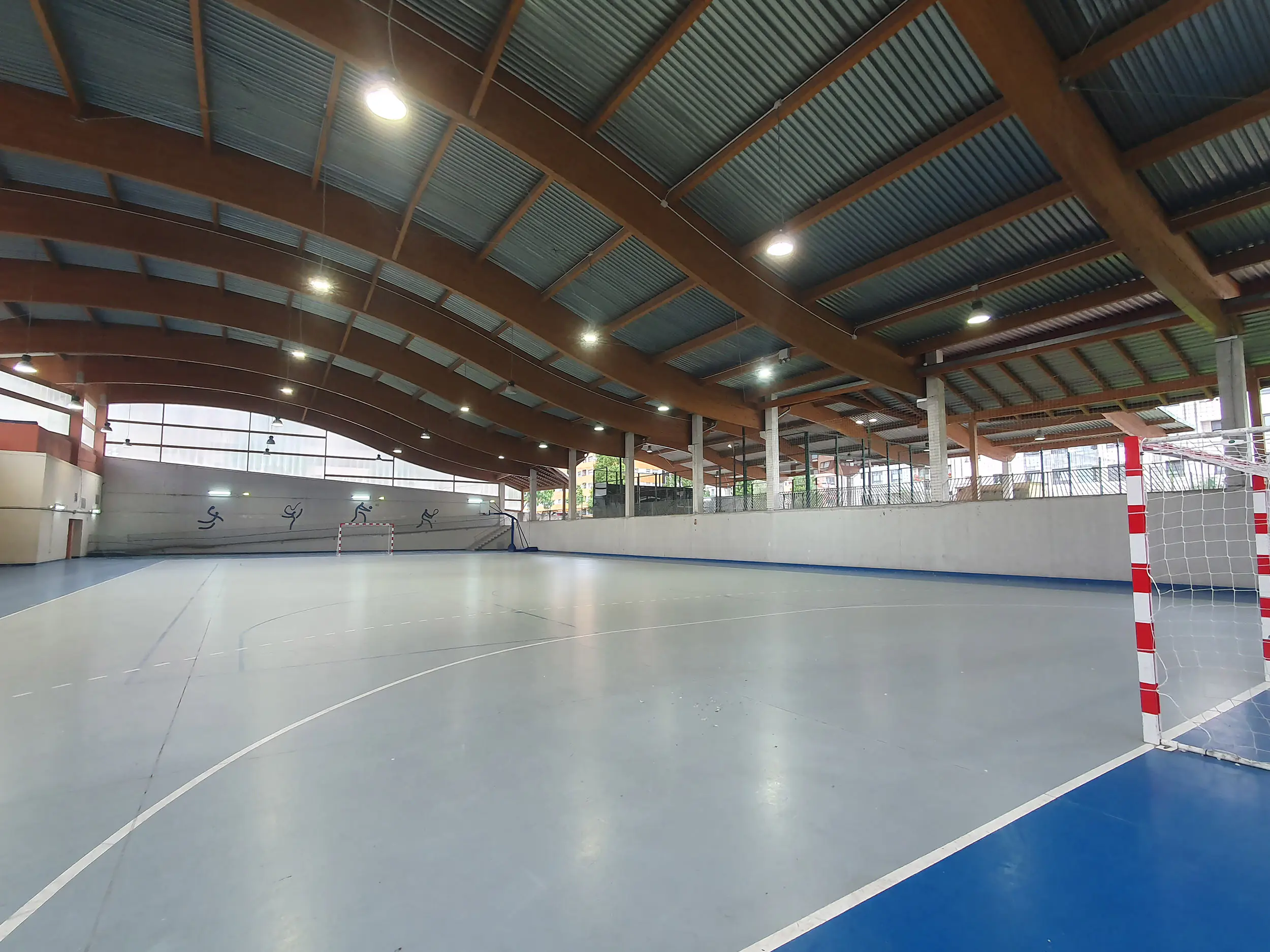 Panorámica desde una esquina de la pista polideportiva, con porterías en los fondos y cubierta ondulada de vigas de madera