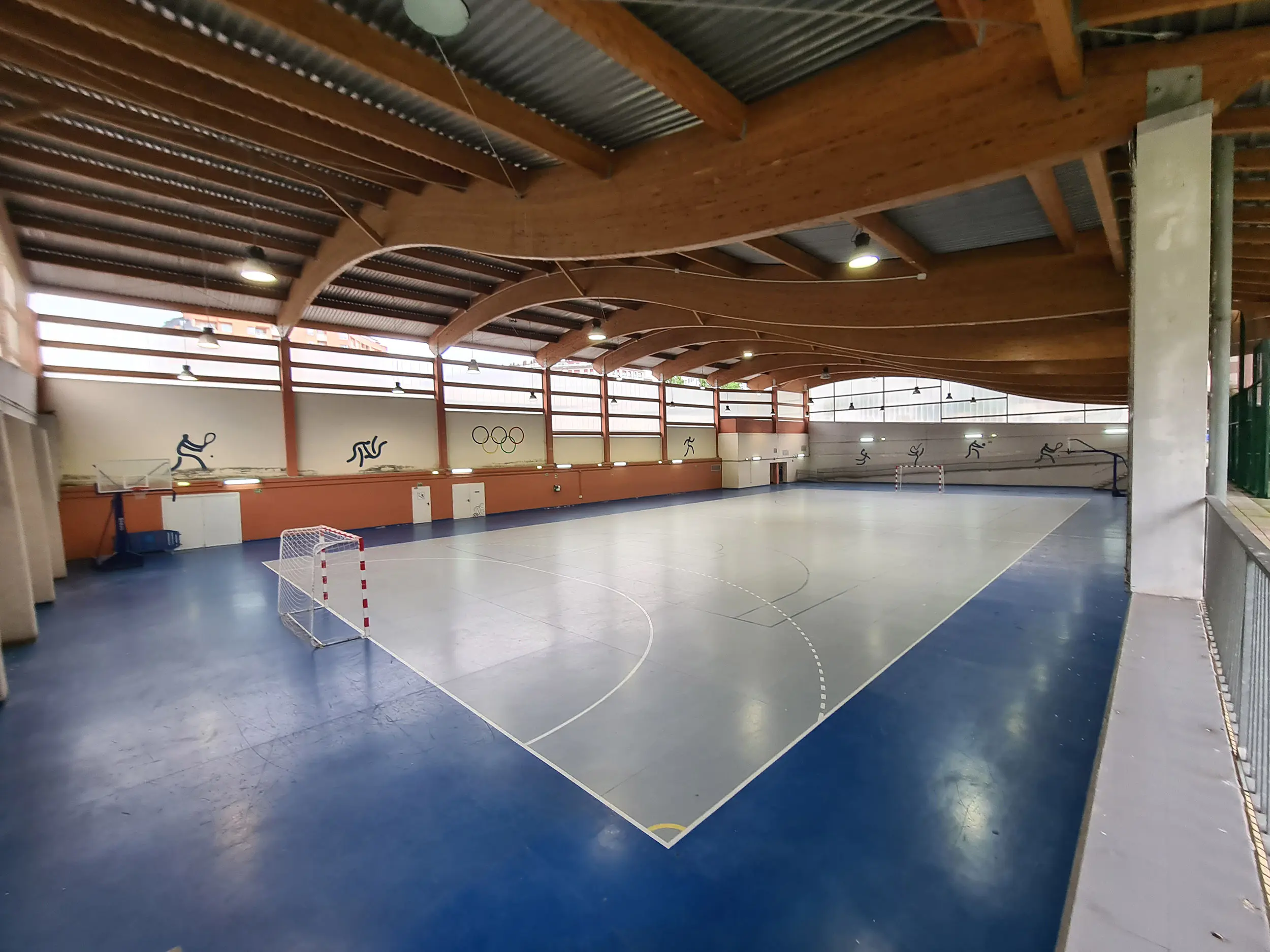 Pista polideportiva desde un ángulo elevado, con porterías, canastas y detalle de la cubierta ondulada de vigas de madera