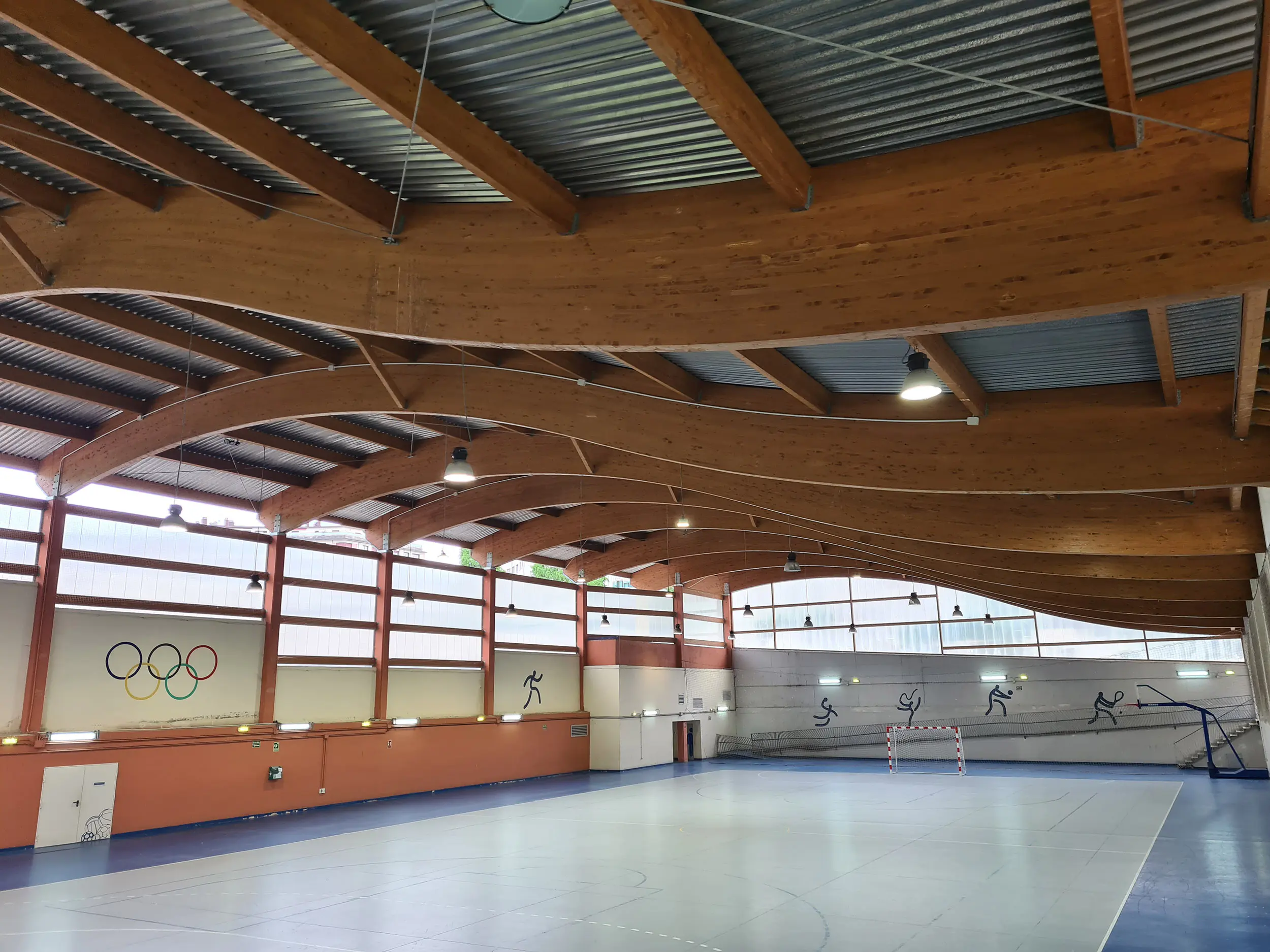 Panorámica de la pista polideportiva, con una portería y una canasta al fondo y una cubierta ondulada de vigas de madera