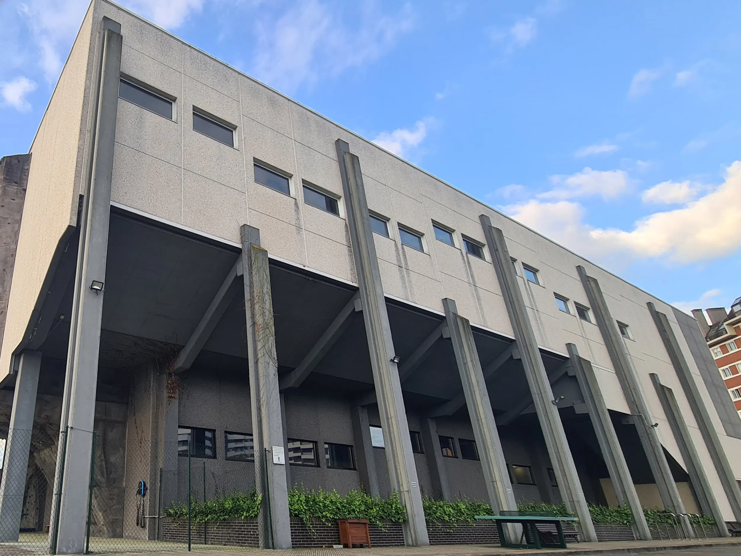 Vista lateral del exterior del edificio, con plantas ornamentales en un pasillo bajo sus columnas y una mesa de ping-pong