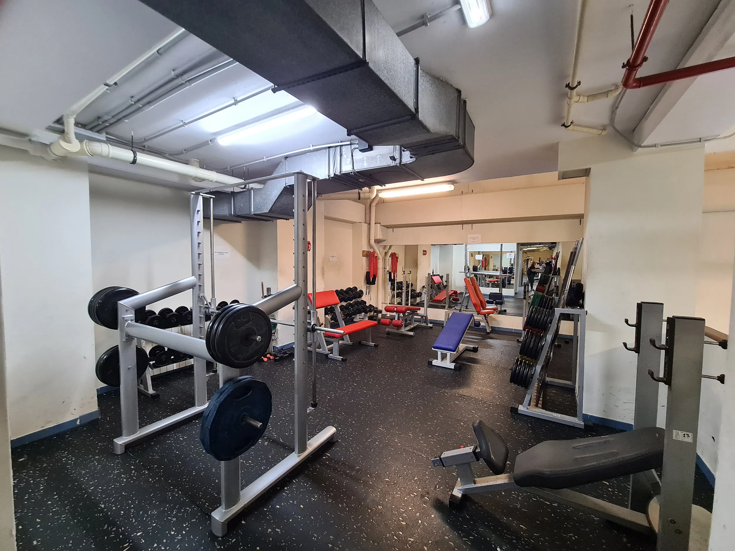 Gimnasio interior, con máquinas de pesas y bancos de musculación, suelo de linóleo y espejo grande en el fondo
