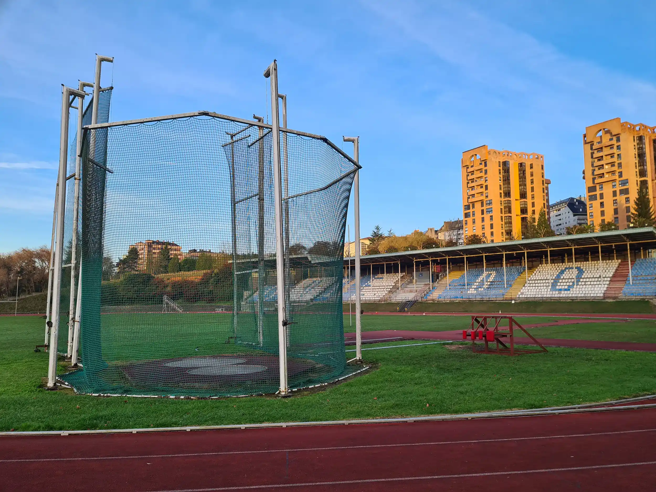 Vista lateral de las pistas de atletismo, con las gradas al fondo y detalle de la jaula de lanzamiento de martillo