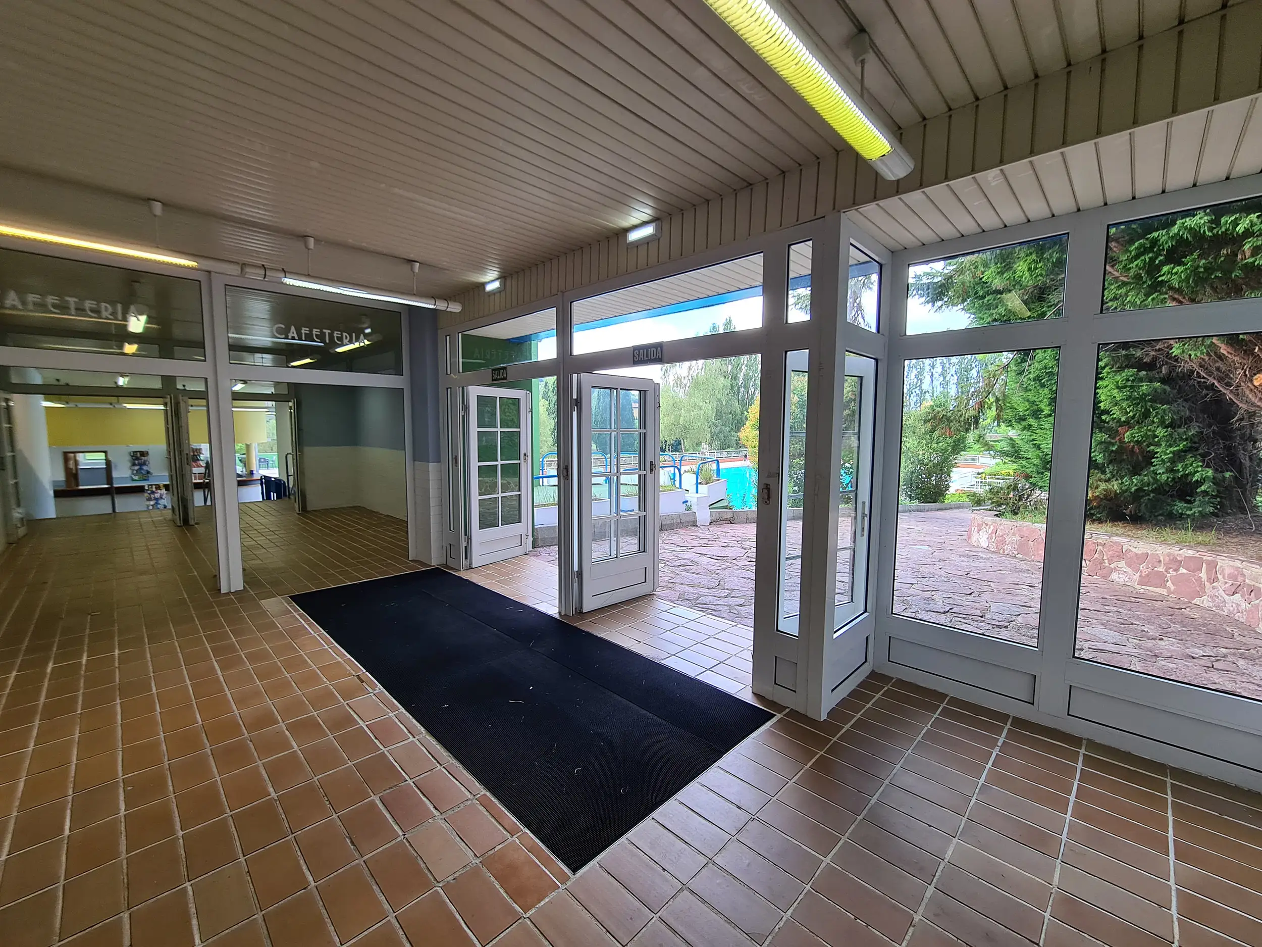 Vestíbulo de distribución, con puerta de acceso a la cafetería y puerta exterior de salida a las piscinas