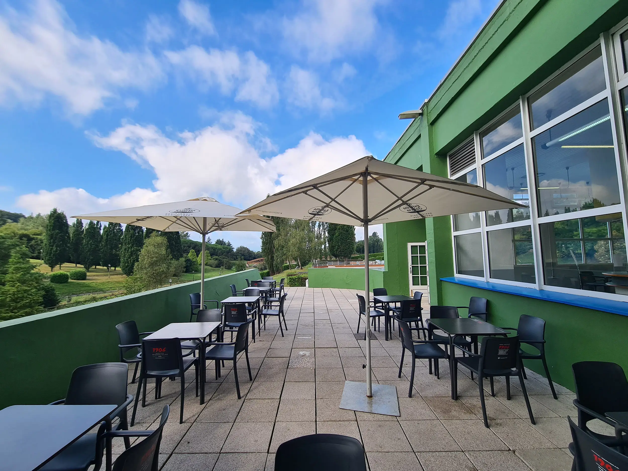 Exterior de la cafetería, con mesas, sillas y sombrillas. Vistas de la piscina principal y el Parque de Invierno