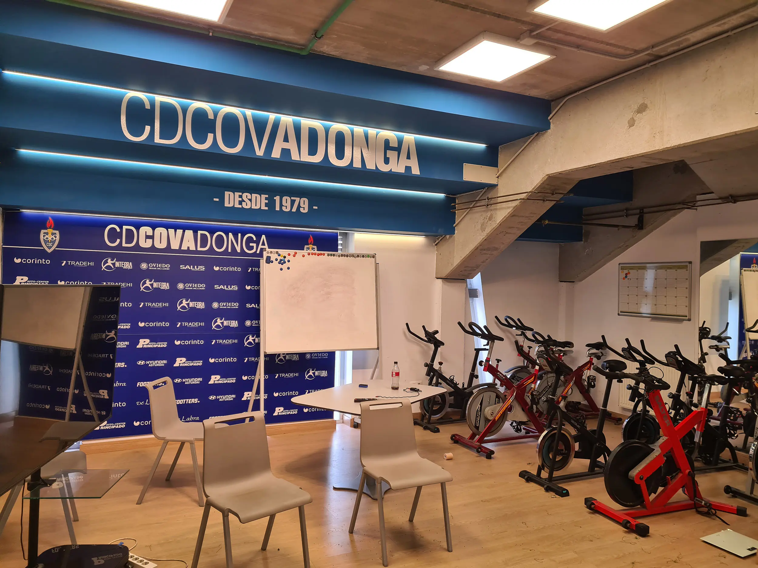 Sala técnica con sillas, mesa y pizarra delante muro con logos de diferentes marcas en letras blancas. Bicicletas estáticas