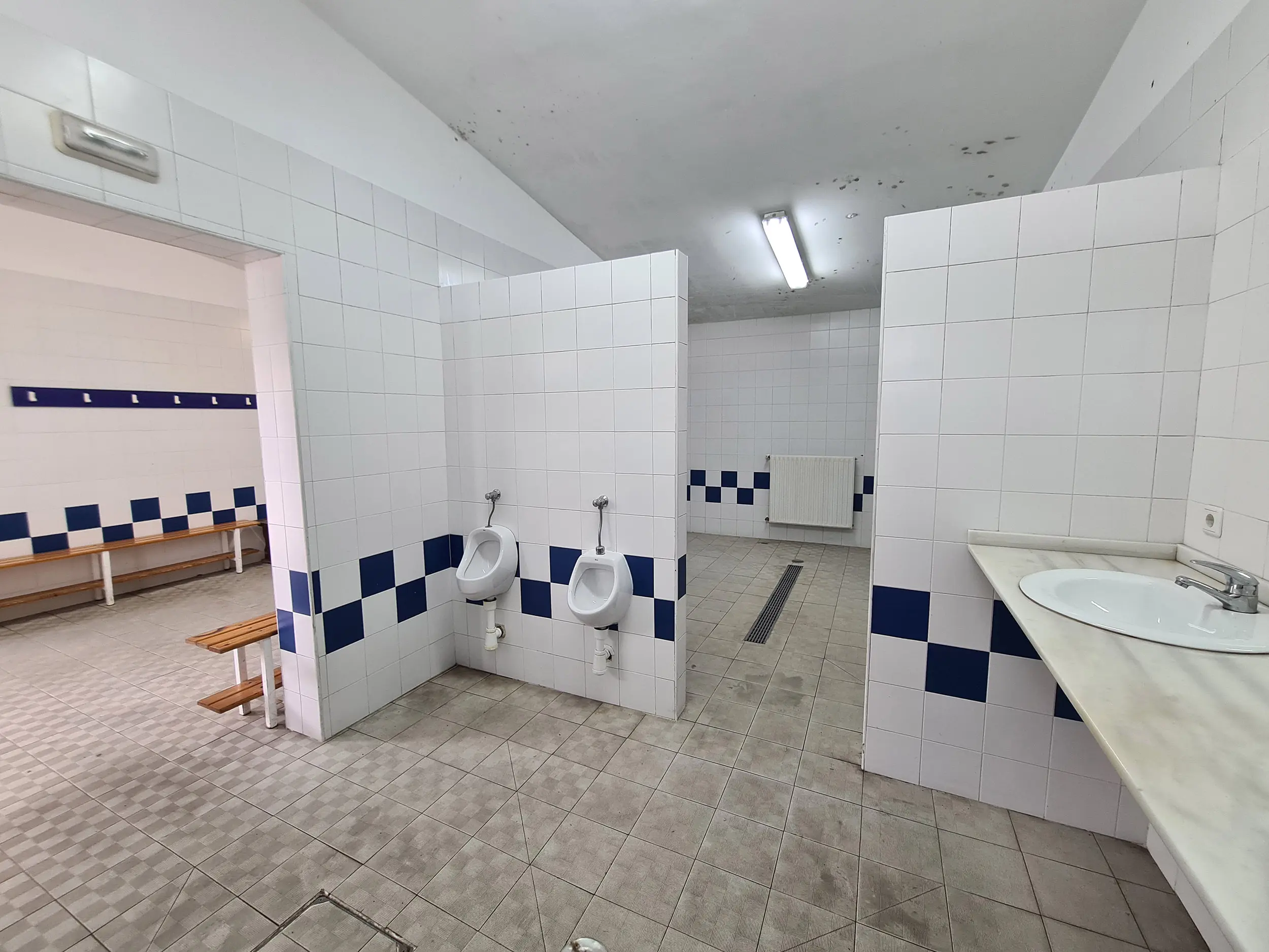 Zona de aseos del vestuario con azulejos blancos y dos hileras de alternos azules y blancos. Además, dos urinarios y lavabo