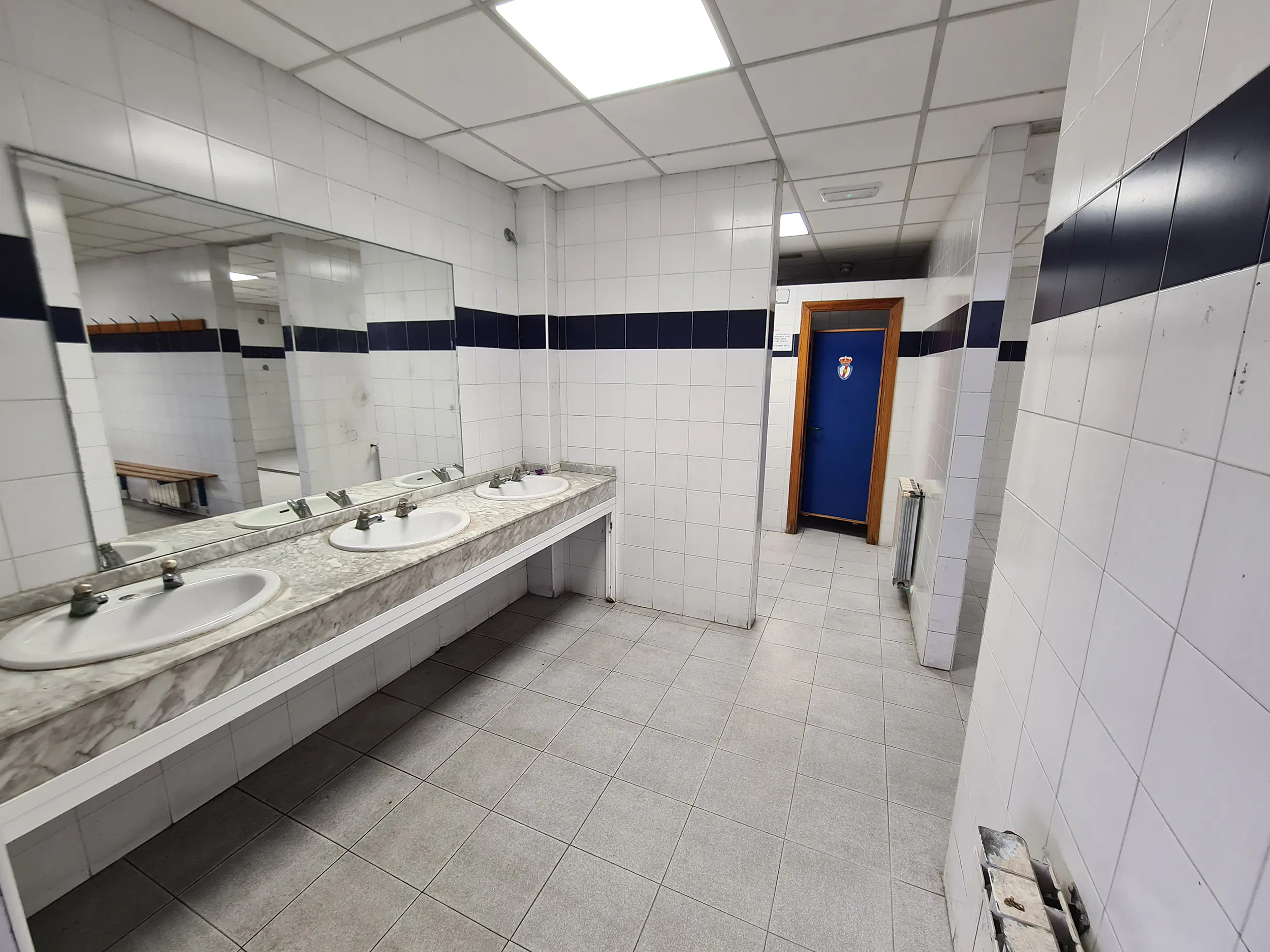 Aseos con suelos y paredes de azulejos, con lavabos y espejos a la izquierda, y al fondo, puerta decorada con un escudo