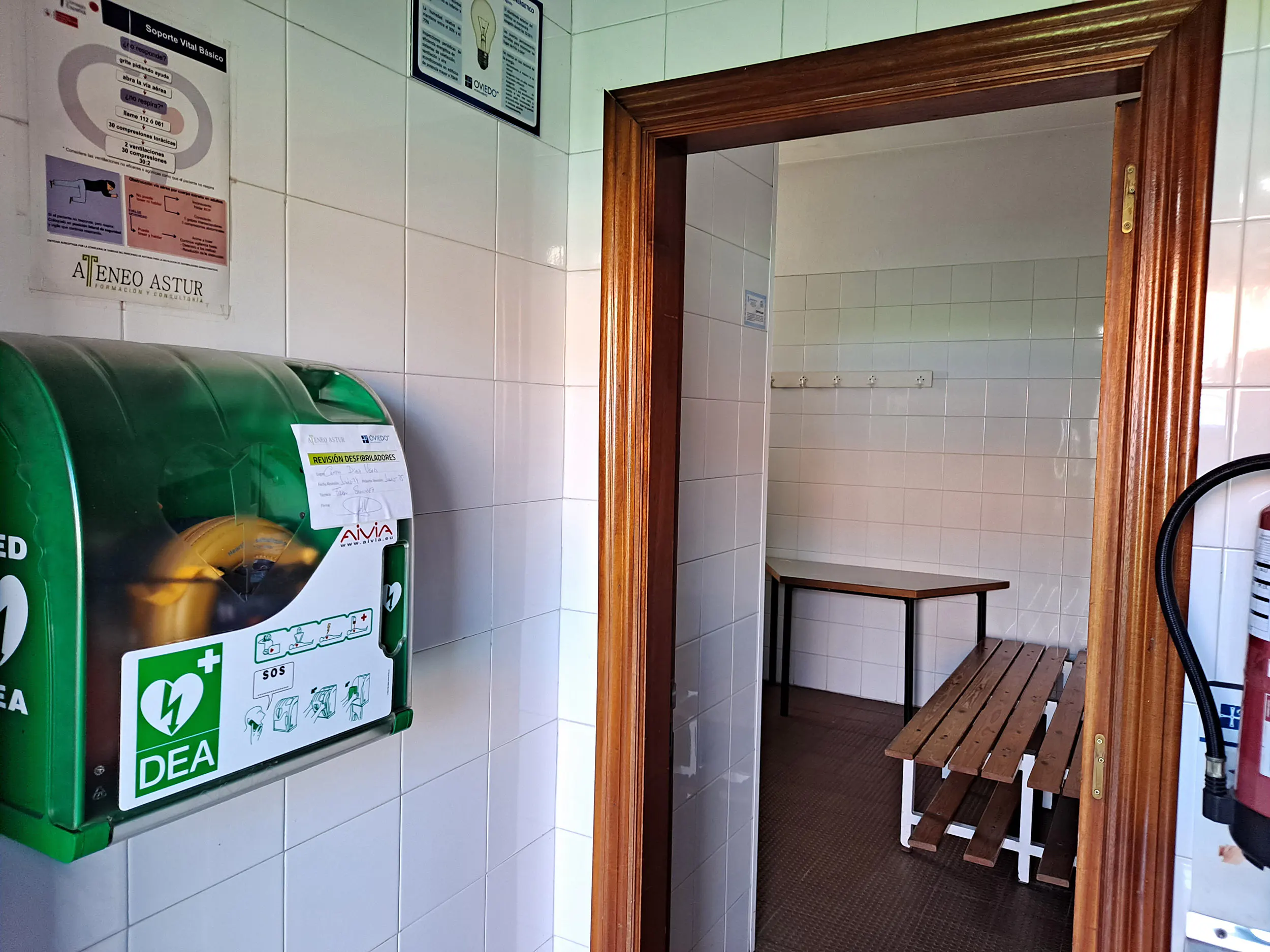 Desfibrilador de pared, junto a la puerta de acceso al vestuario, con bancos de madera en su interior