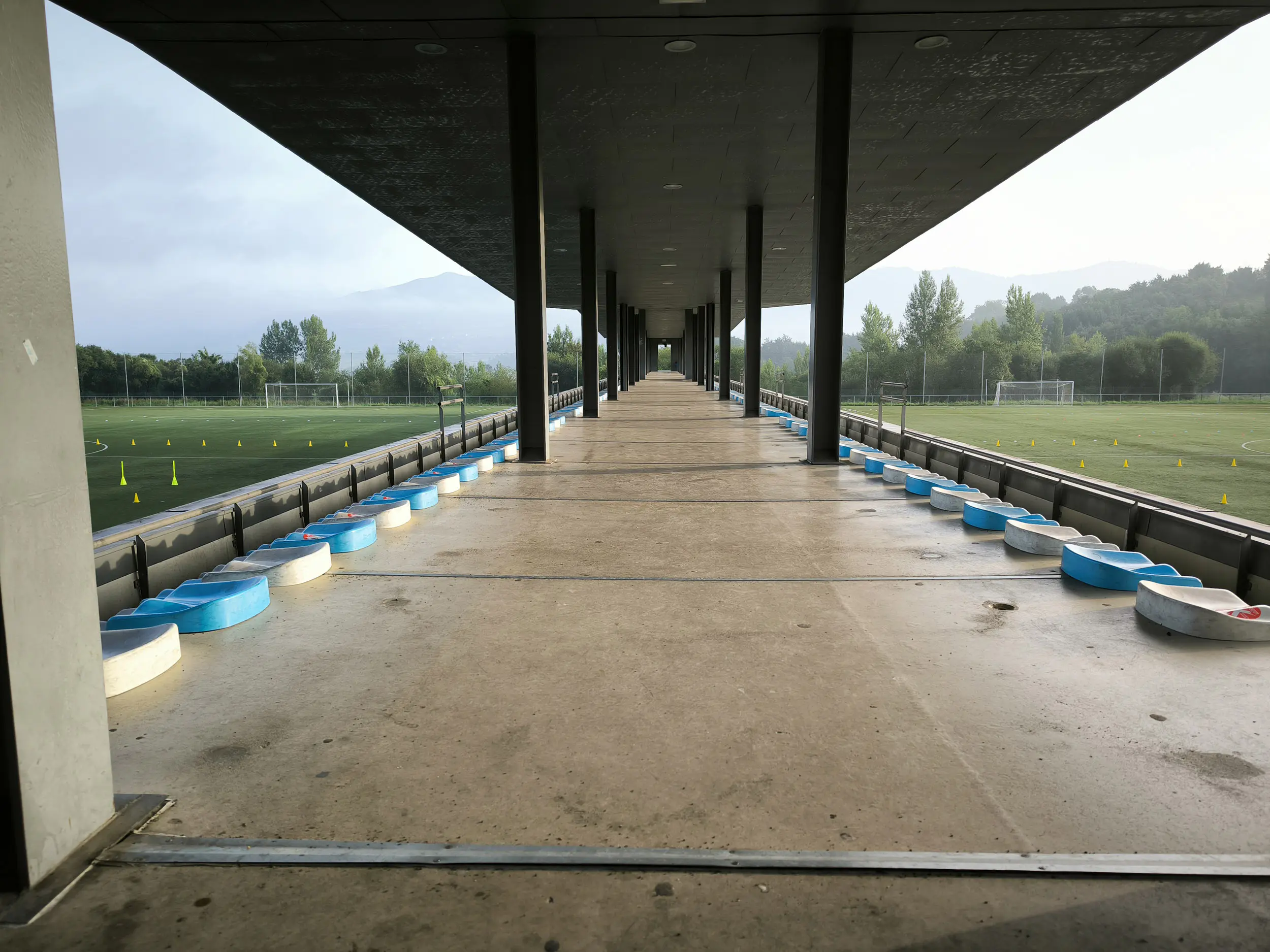Pasillo cubierto de acceso a las gradas, con columnas y vista parcial de los dos campos de fútbol 11 a los lados
