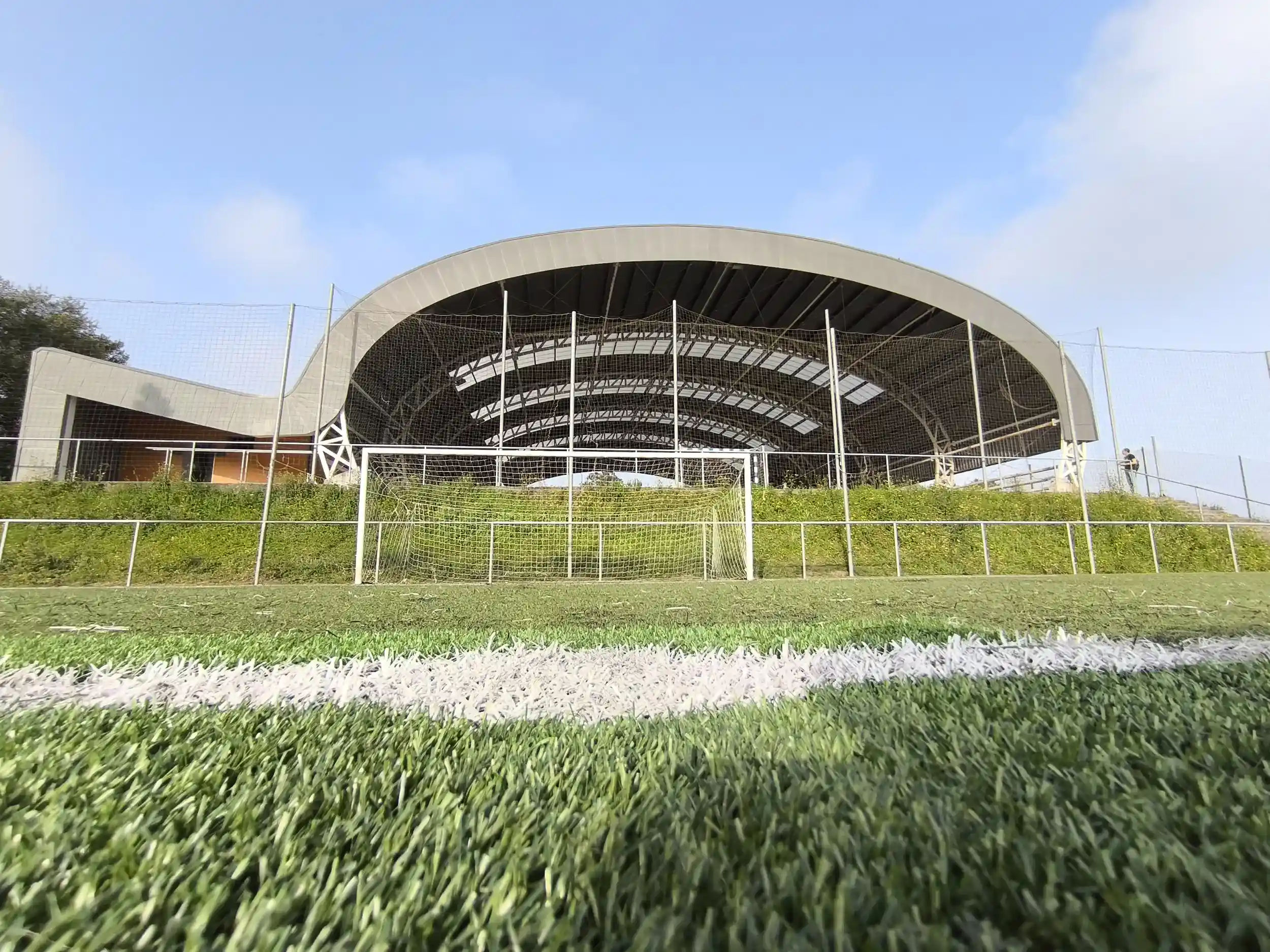Vista parcial, desde el nivel del suelo, de uno de los campos de fútbol 8, con la cubierta de la pista polideportiva al fondo