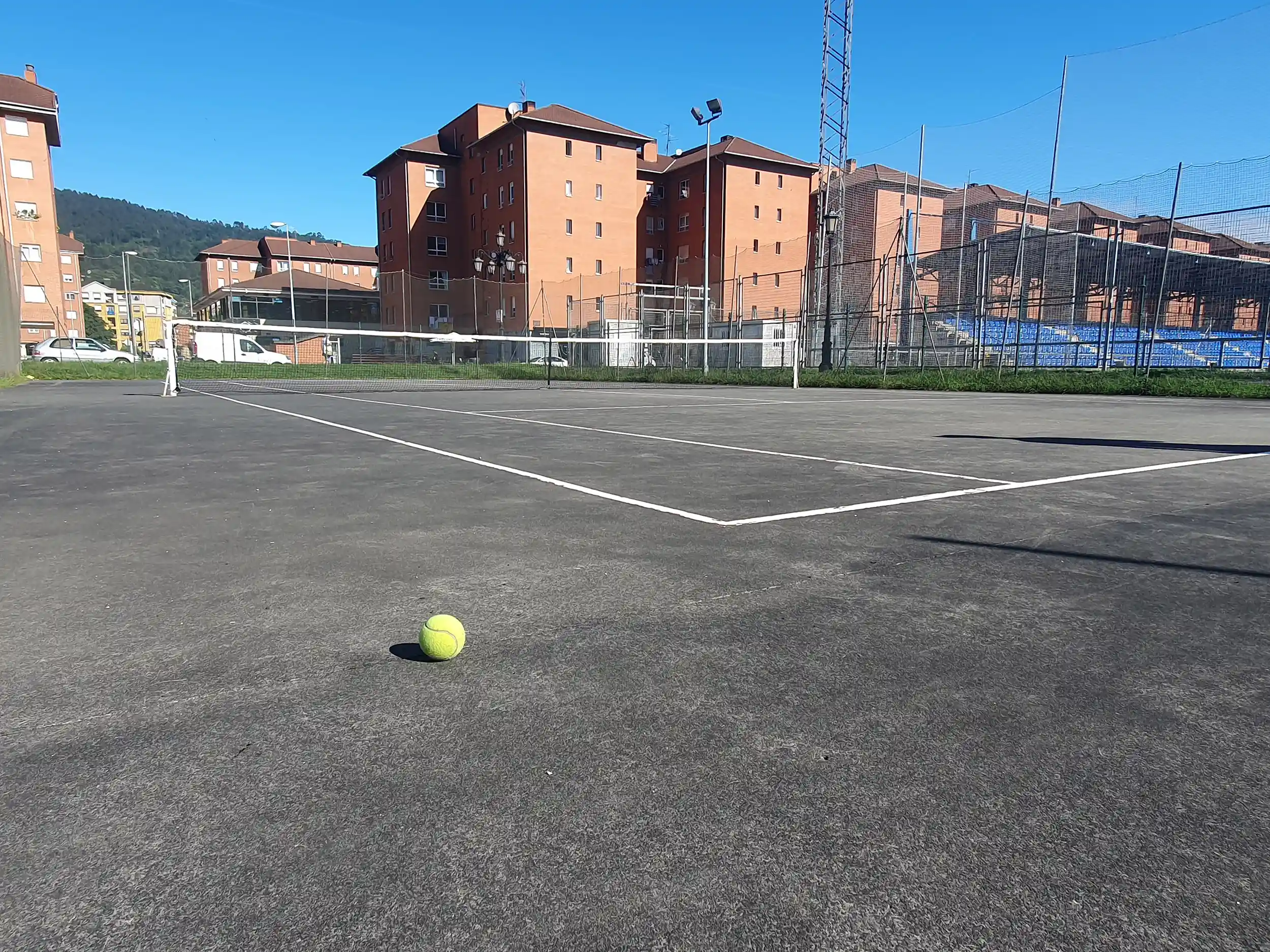 Vista lateral de una de las pistas de tenis, con una pelota en primer término y edificios de viviendas al fondo