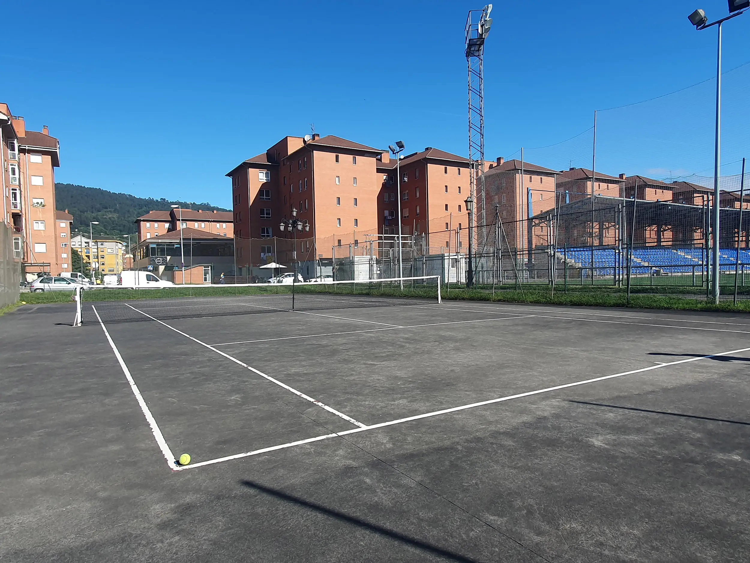 Vista lateral de una de las pistas de tenis, con una pelota en primer término y edificios de viviendas al fondo