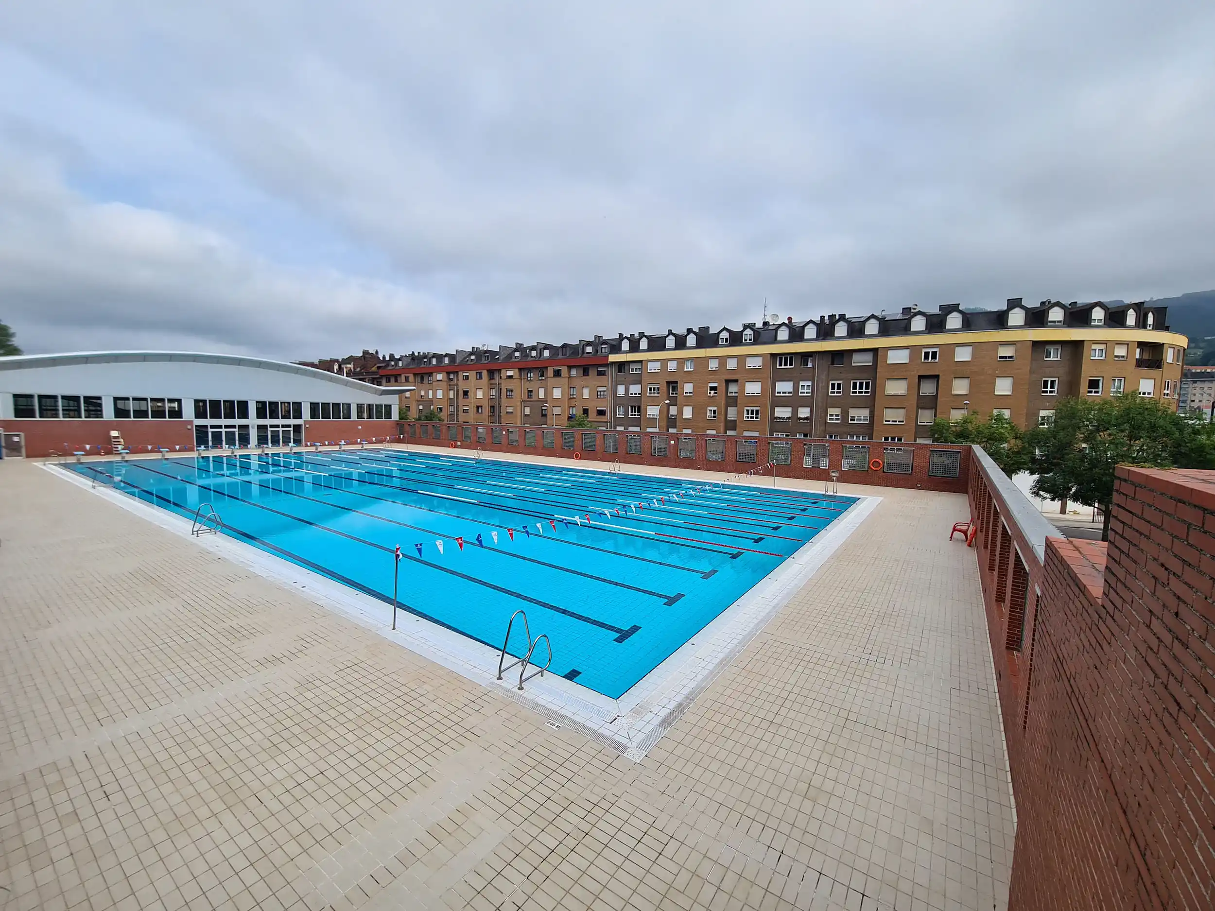 vista elevada de la piscina desde una esquina. A la izquierda el edificio de las piscinas cubiertas