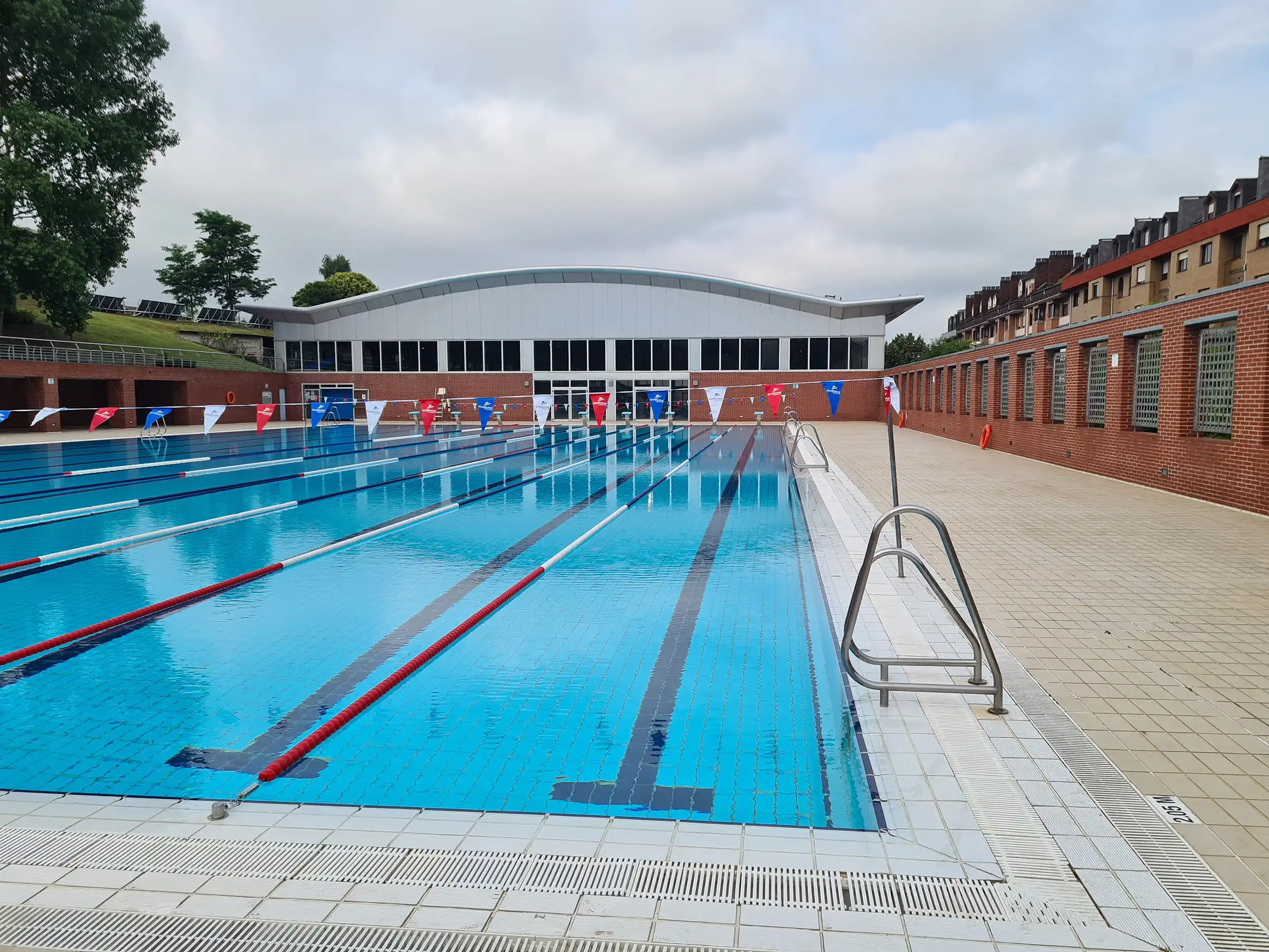 vista parcial de la piscina, en primer plano de una de las escalerillas y, al fondo, edificio de piscinas cubiertas 
