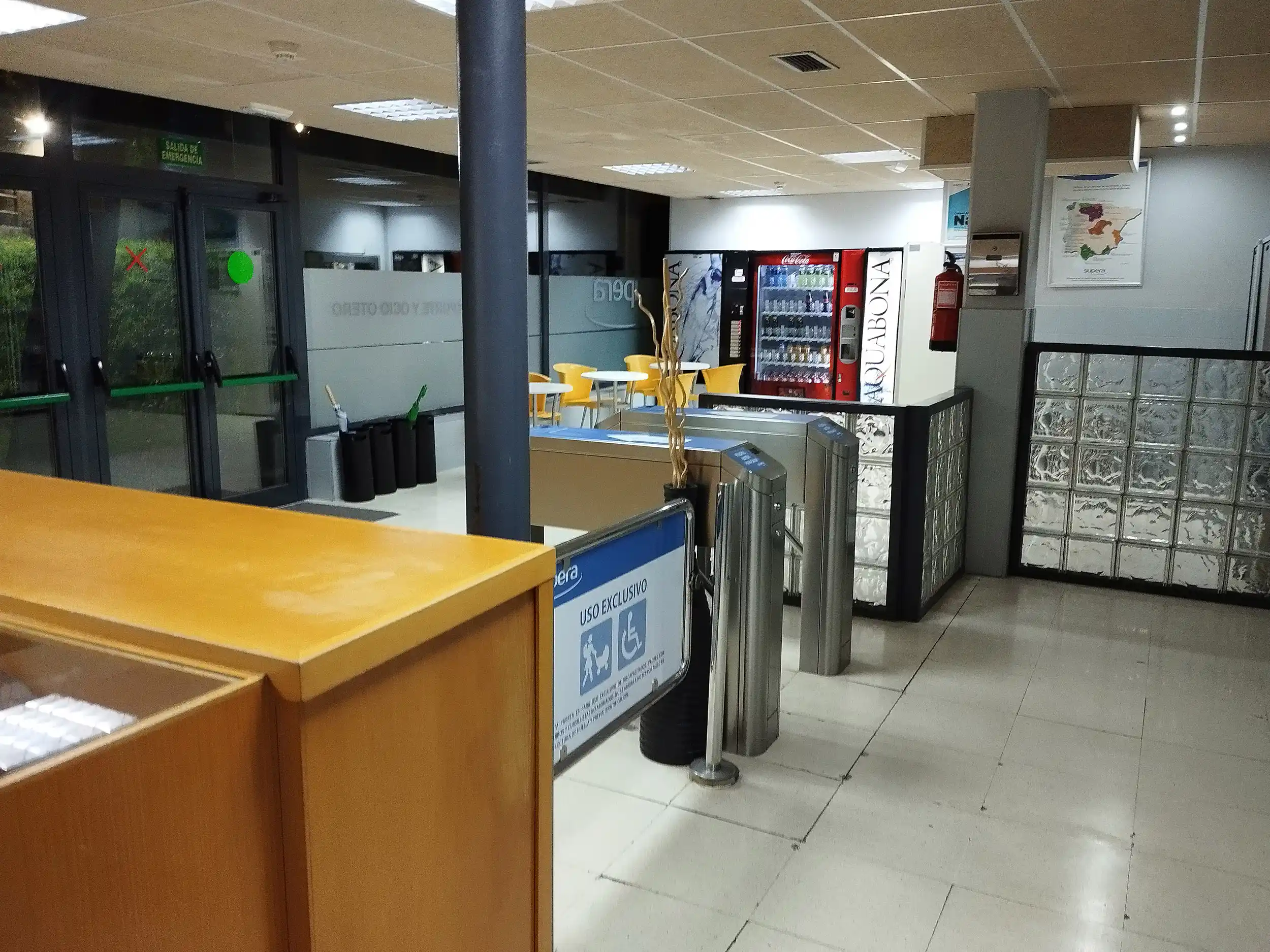 Acceso a las instalaciones, con tornos de acceso, mostrador de recepción y, al fondo, mesas, sillas y máquina expendedora