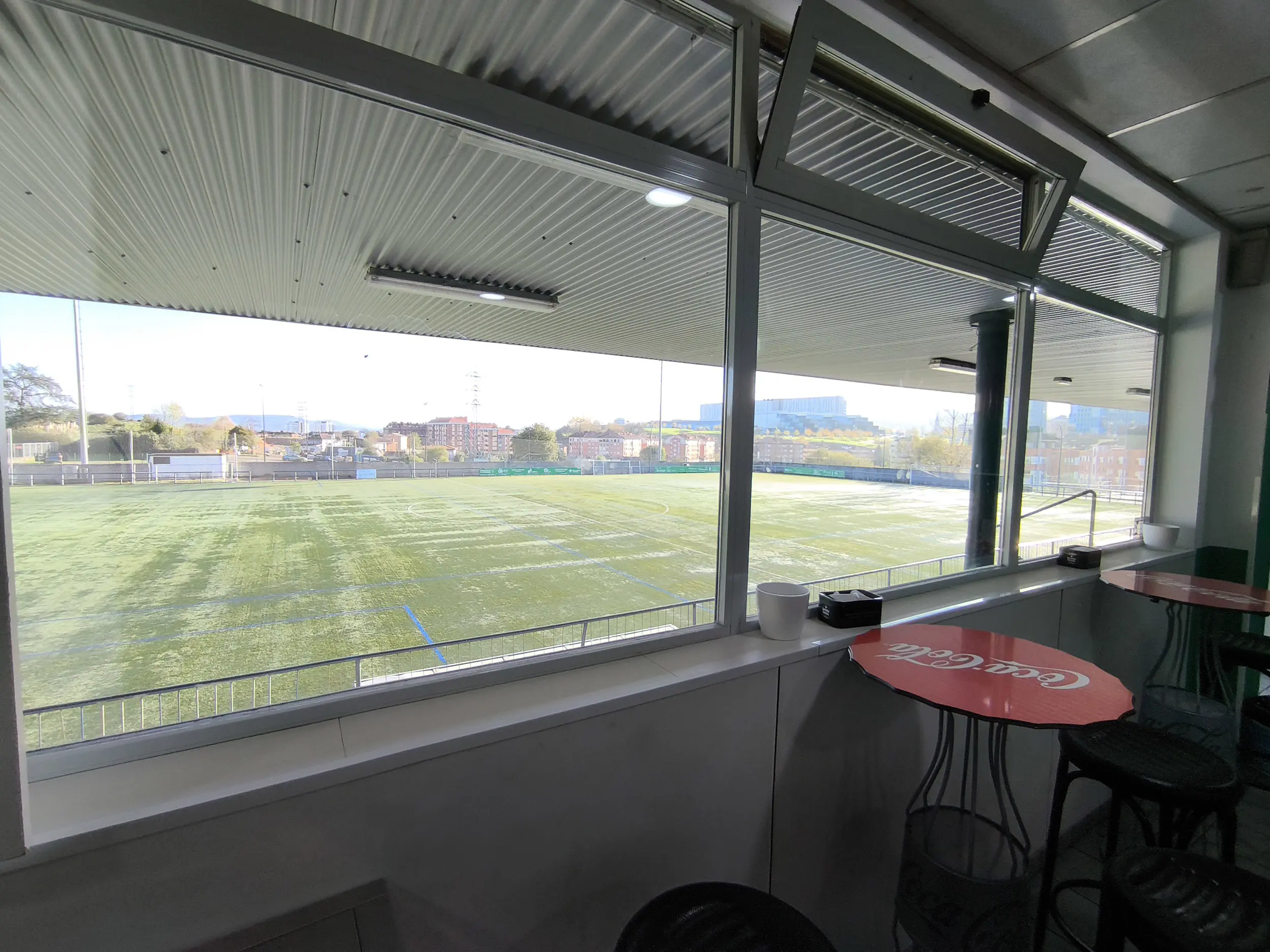 Campo de fútbol a través de los ventanales de la cafetería con mesas altas rojas con taburetes
