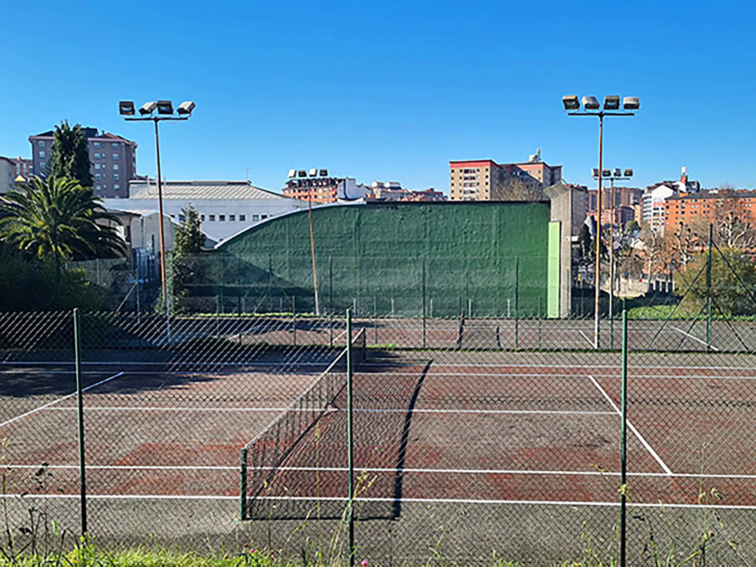 Vista lateral de una pista de tenis, con la red central, vallas, torres de iluminación y, al fondo, la pared de un frontón