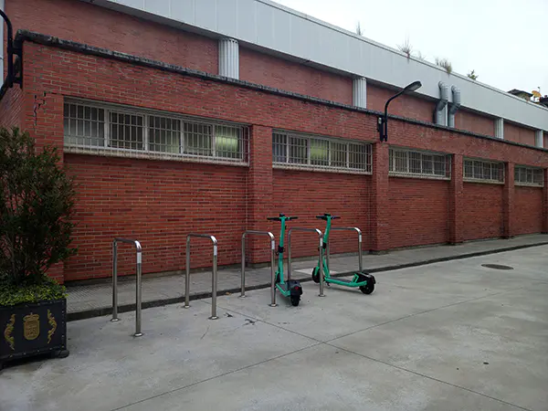 Aparcamiento de bicicletas al lado del muro lateral del edificio polideportivo. Aparecen 2 patinetes eléctricos