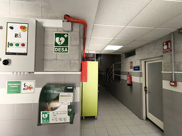 Pasillo de acceso a vestuarios y planta superior. Armario de maniobras eléctricas y aparato desfibrilador semiautomático