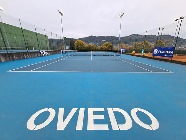 Vista de una cancha de tenis de green set descubierta, con el texto Oviedo pintado en la pista, en primer plano