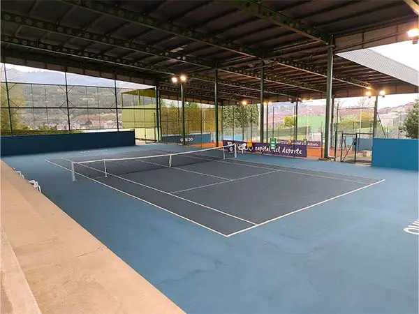 Panorámica de una pista de tenis cubierta desde la grada lateral, con vista de la cubierta y las vallas perimetrales