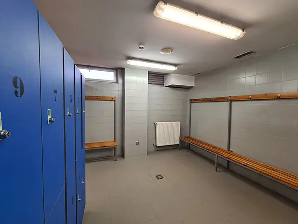 Interior de los vestuarios, con bancos, perchas adosadas a la pared y taquillas a la izquierda