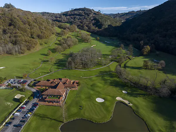 Vista aérea del edificio principal, aparcamiento anexo, un pequeño lago y primeros hoyos del recorrido del campo