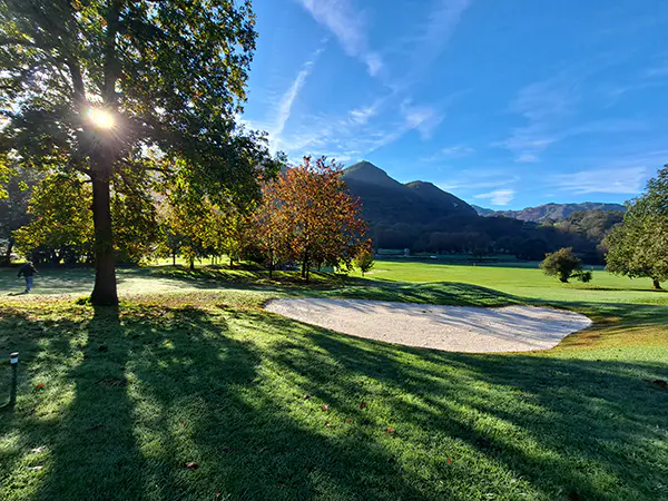 Panorámica de una zona del campo de golf, con un búnker de arena, árboles y un fondo montañoso