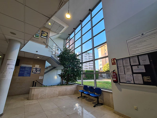 Vestíbulo de entrada, con grandes ventanales, escaleras, asientos y un tablón de anuncios en la pared