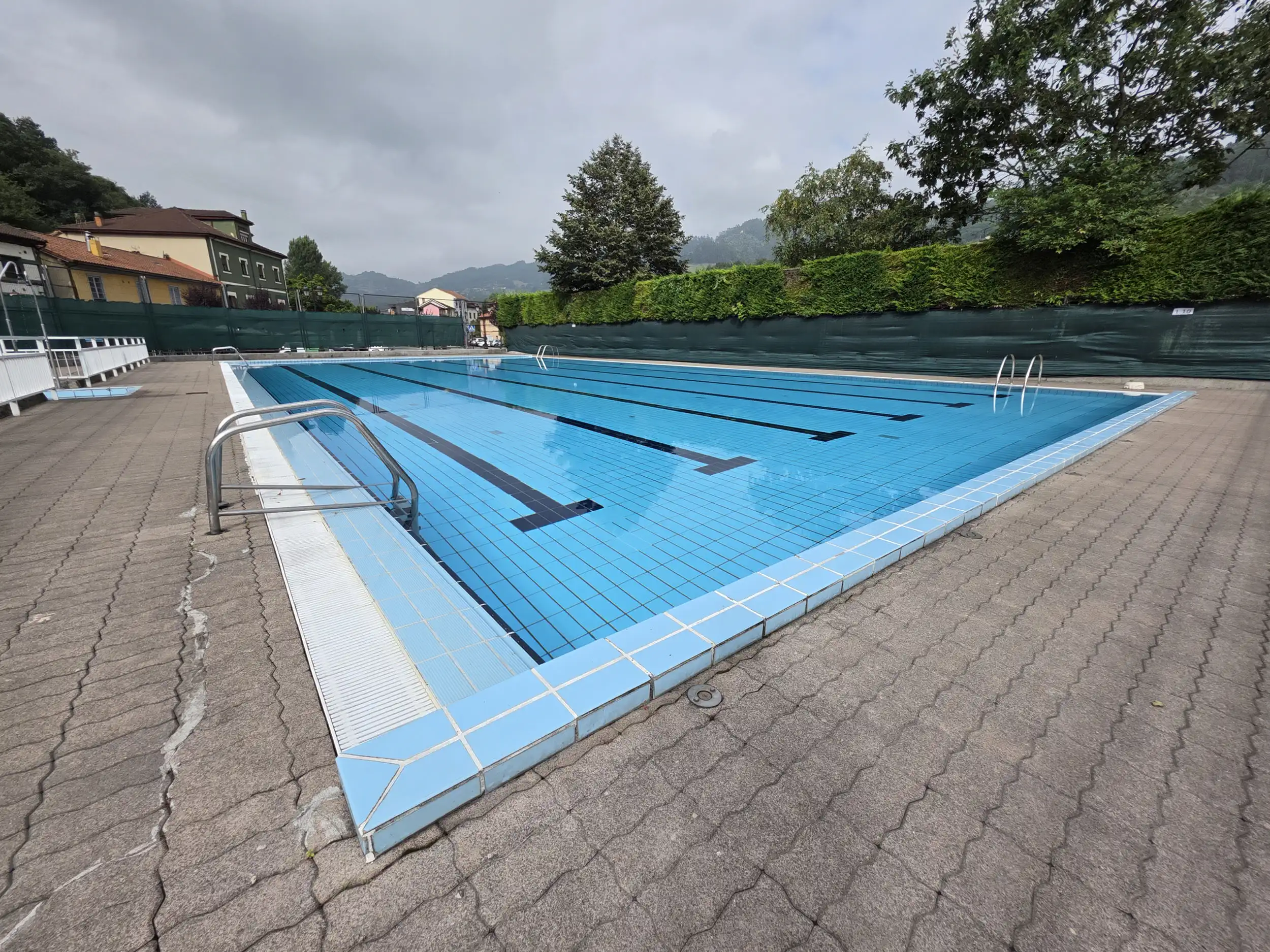 vista de la piscina principal desde una esquina con árboles, arbustos y viviendas al dondo